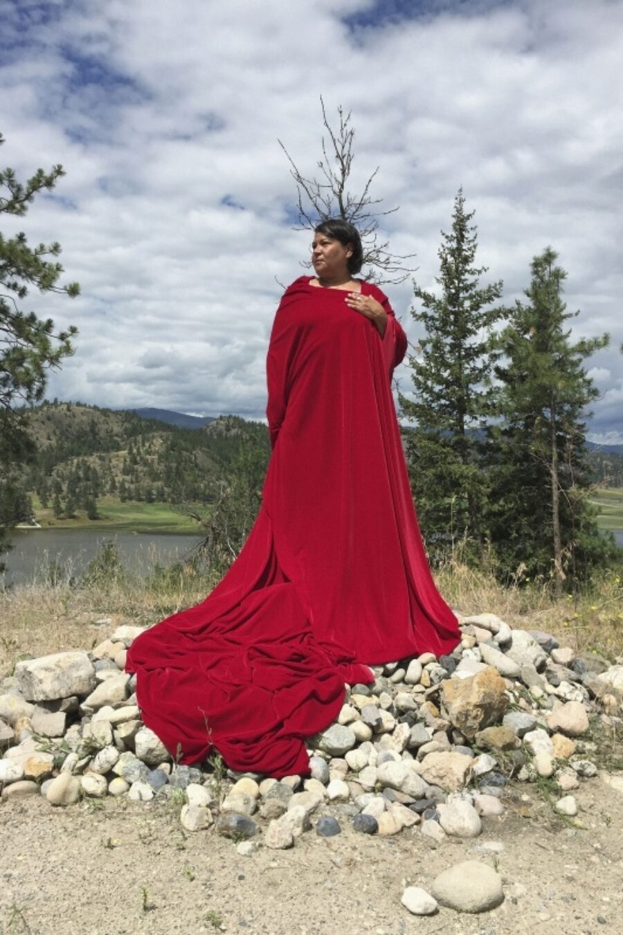 Lori Blondeau stands confidently on a pile of rock in a landscape while wearing a long red dress.