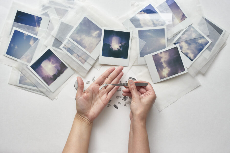 A number of Polaroids lie on a white table while hands pick up paper fragments with tweezers.