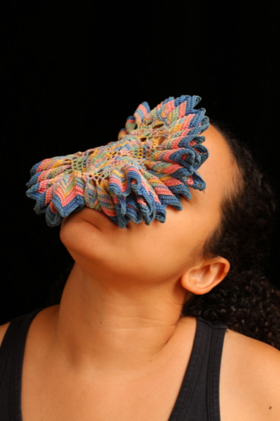 A circular, multicoloured crocheted piece sits on a person's face while they tip back their head.