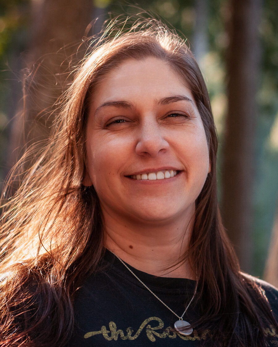 This is a headshot of Jenel Shaw, a white woman in her early 40s with long straight brown hair. Jenel is smiling and squinting at the viewer and is wearing a black T-shirt with yellow writing and a silver necklace.