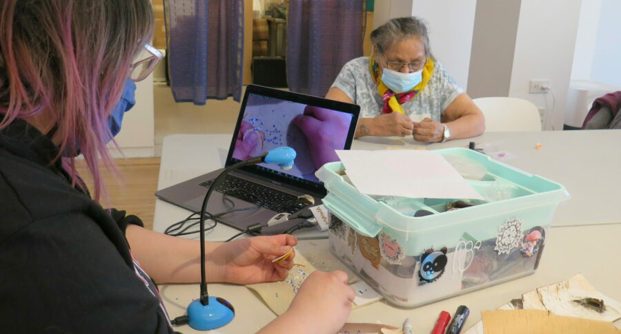 A photo of two people working on beading projects at a table. One person is using a laptop and webcam to share their work over Zoom.