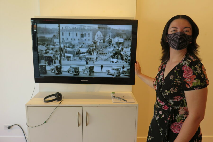 Sarah Paradis in a floral print dress and wearing a black mask, stands next to a television showing a black and white archival image of Winnipeg.