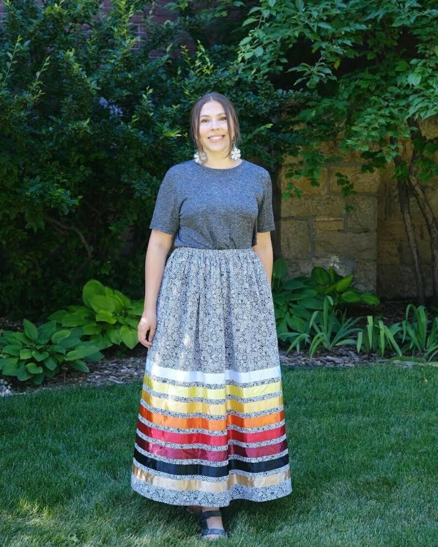 Photo of Zoe Stevens. Stevens stands outside on a lawn and is wearing a ribbon skirt.