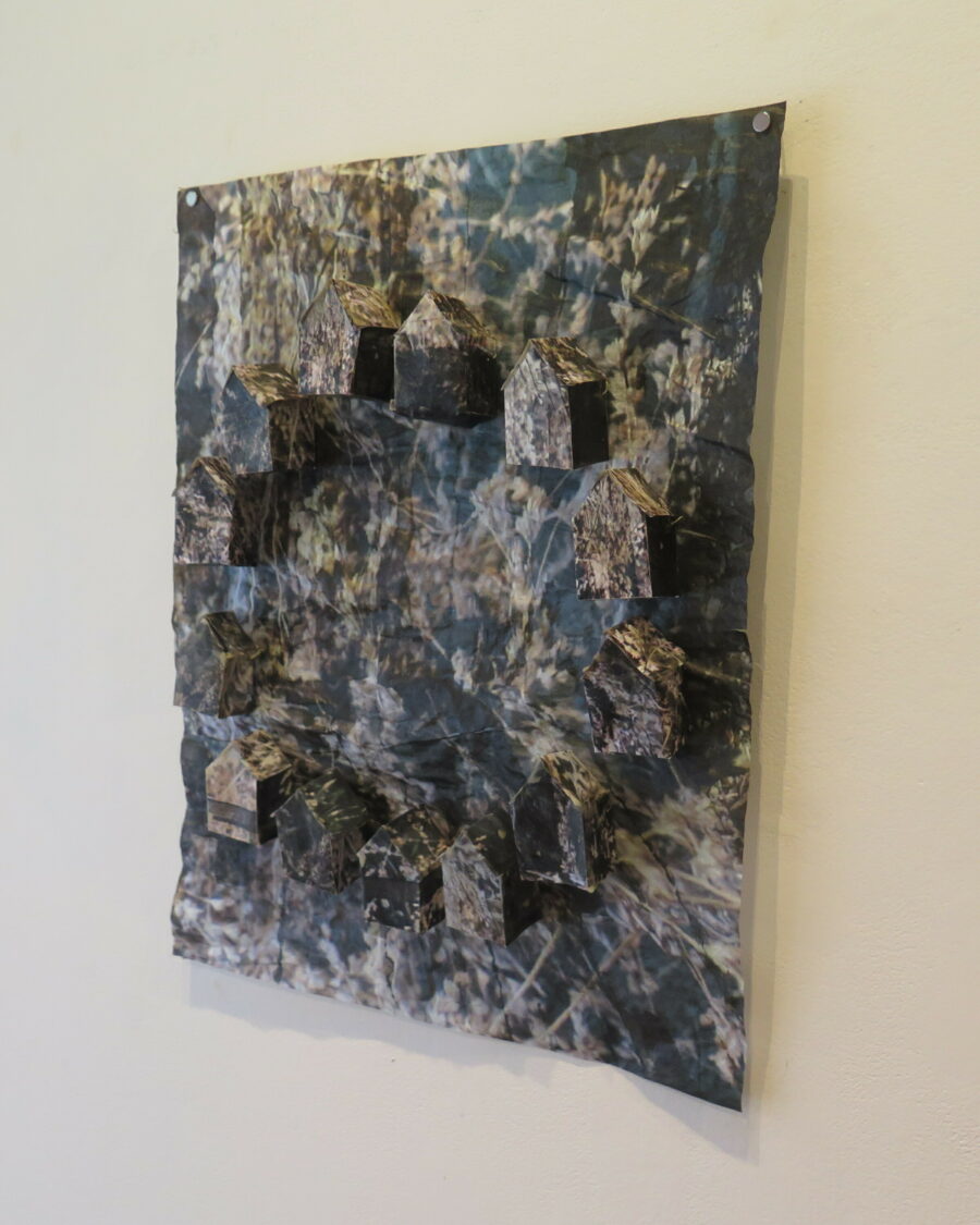 Photo of the portion of the artwork on the wall. It is a piece of paper with a photograph of earth and grasses printed on it.  12 3-d houses made out of the same paper are attached to the paper in a circle.