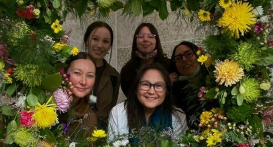The members of the Mîcêt Tipiskâwi-pîsimak Collective, Frances Cooper, Annie Courchene , Tess Ray Houston, Lita Fontaine and Kristy Janvier, look through the center of a very large lush flower wreath.