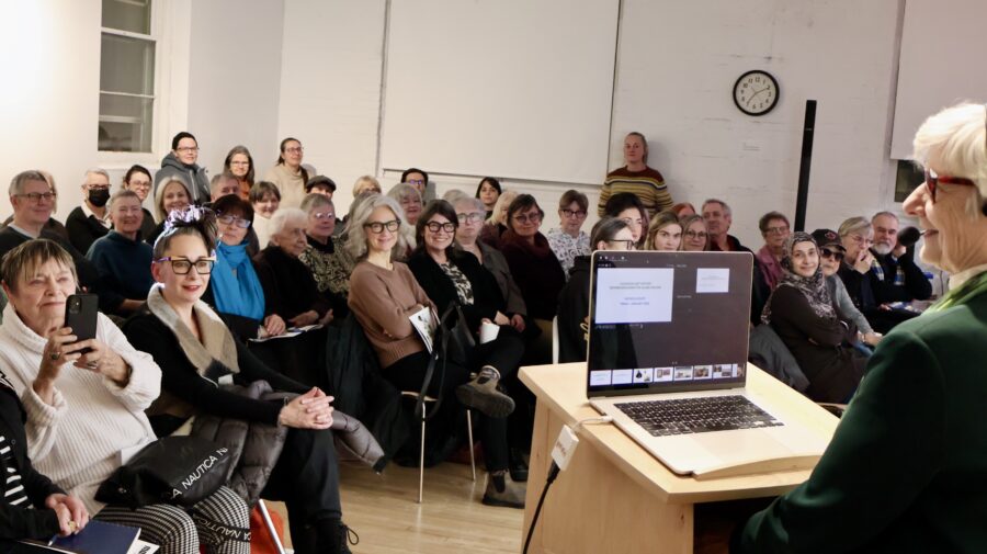 Patricia Bovey stands at a lectern in front of a crowd of 60 people seated in MAWA.