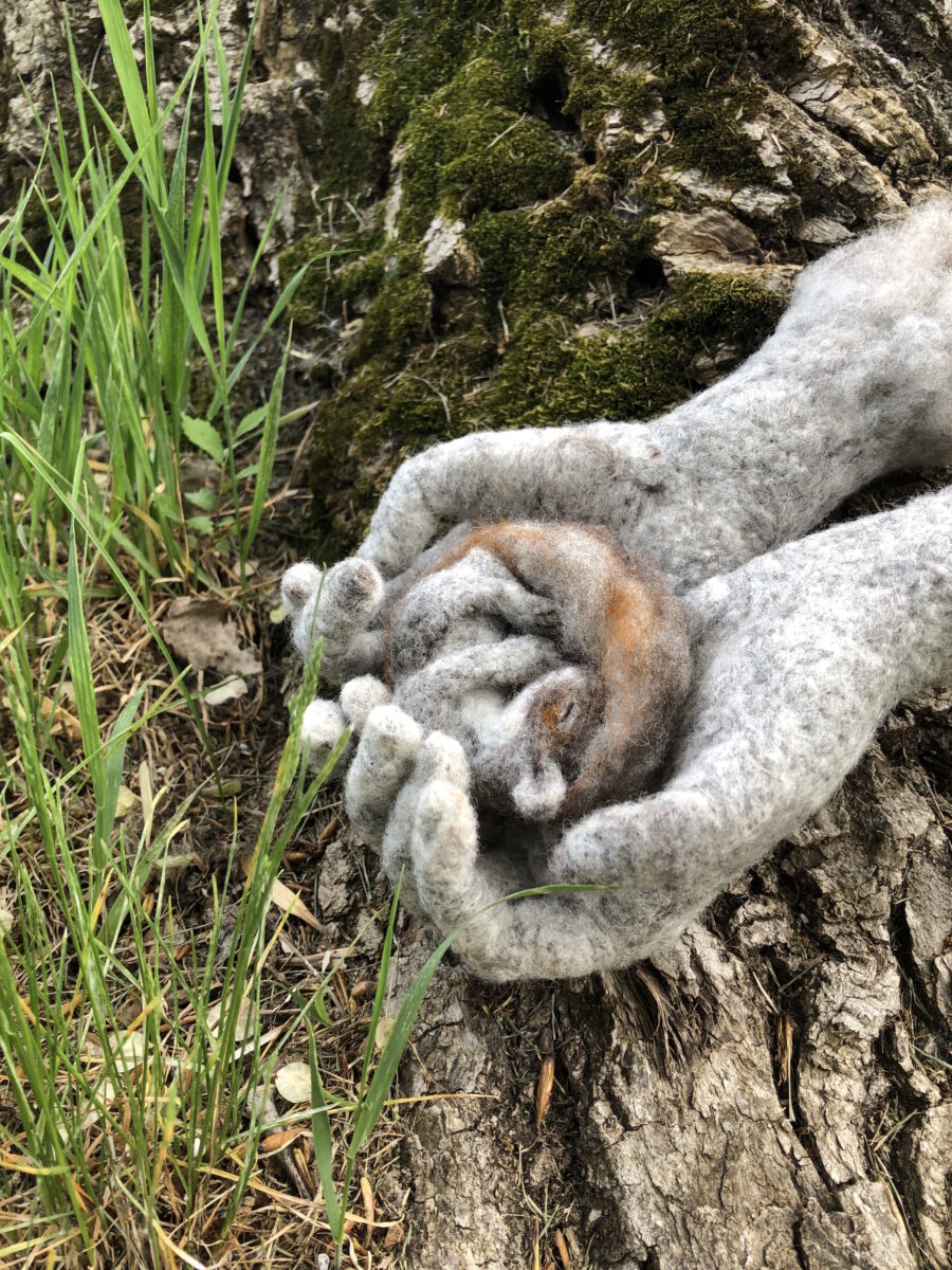 Needle-felted sculpture by Rosemarie Péloquin of two hands cupping a small curled up creature. Pictured outdoors.