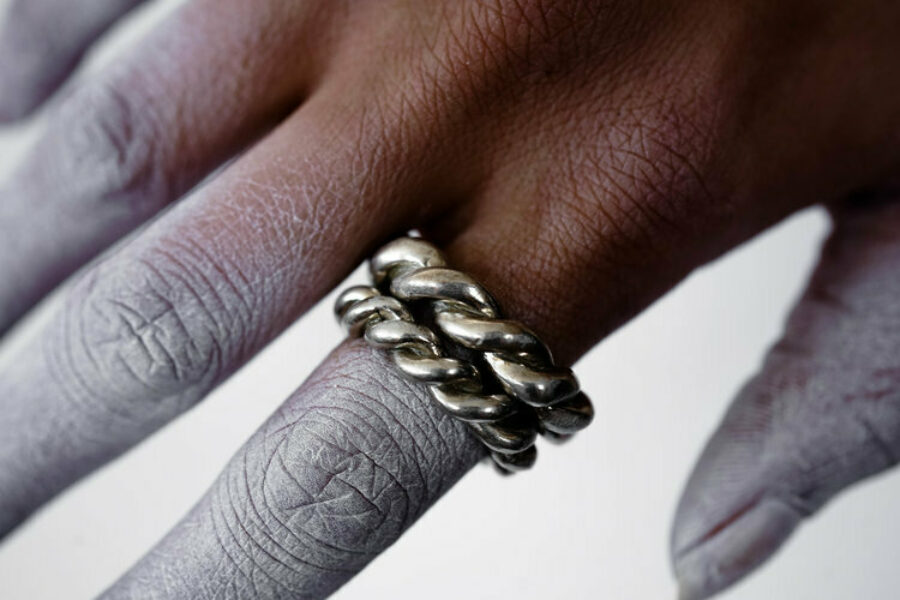 Sadé, helios love, available in a variety of metals. A hand with brown skin and a white substance on the fingers wears a metal ring.