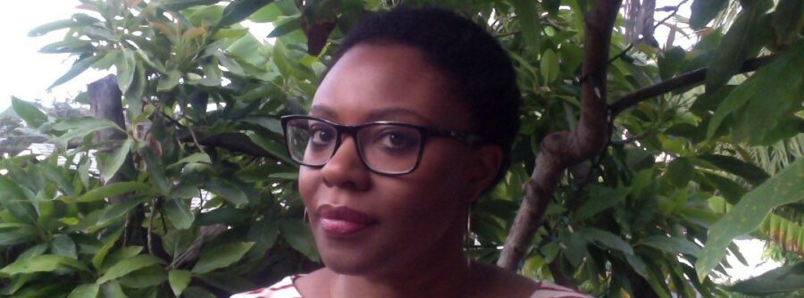 Picture of Sally Frater standing in front of a leafy bush looking directly at the camera. Sally has dark brown skin, short black hair and is wearing glasses.