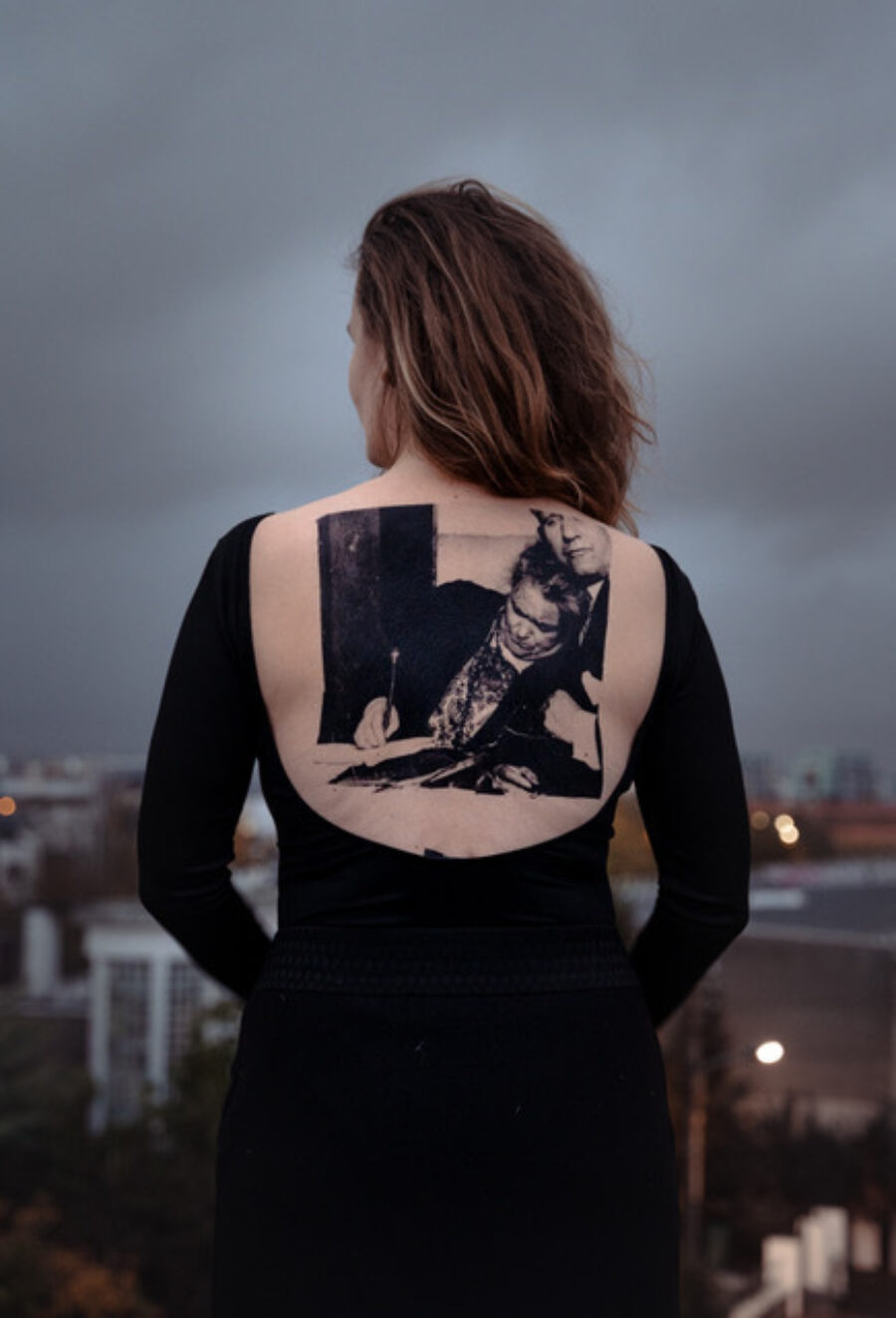 A woman stands with her back to the camera wearing a black shirt that leaves her back bare. An image is imprinted on her skin.