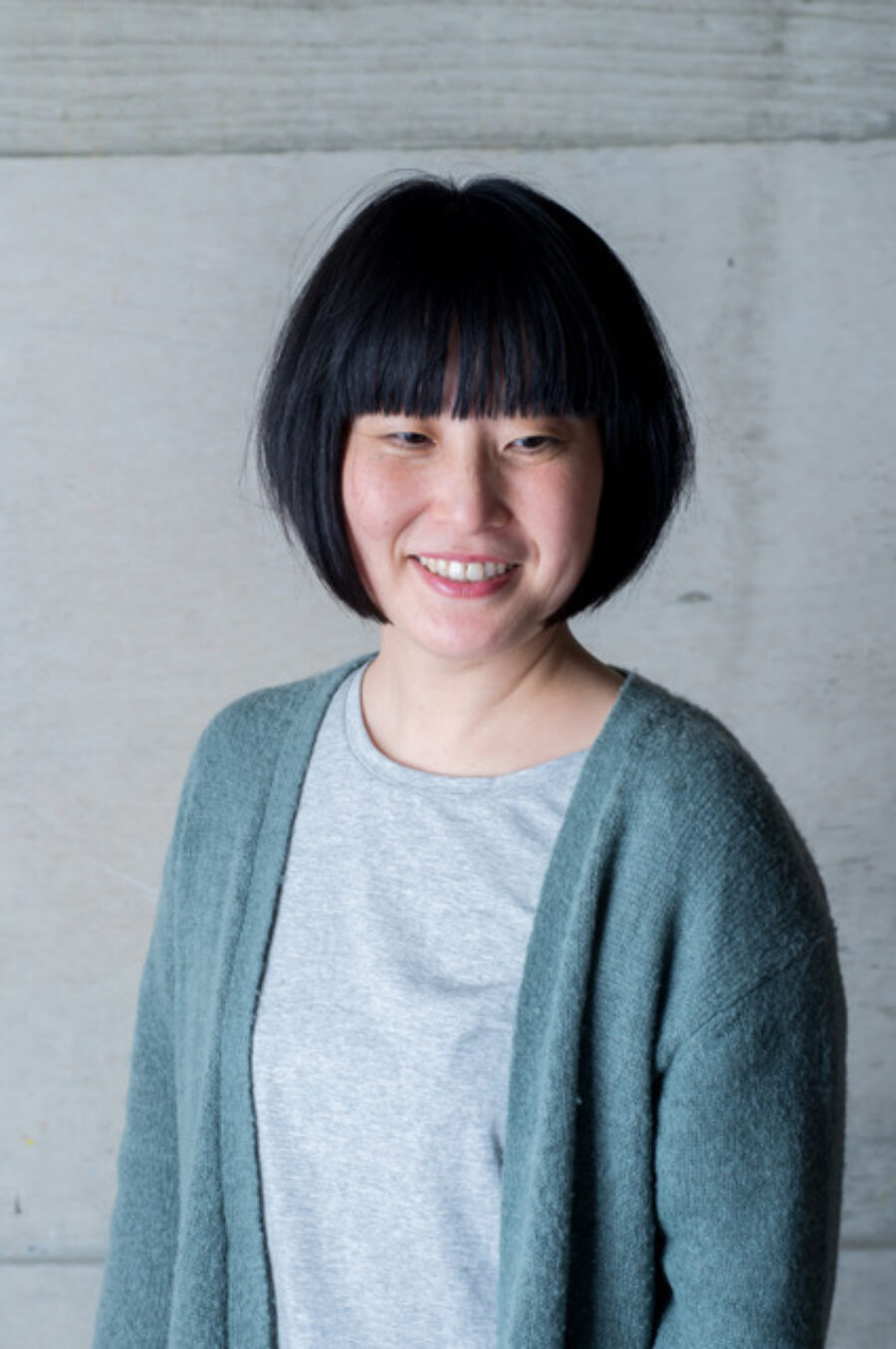 Photo of Tomoko Inagaki standing in front of a white wall. Inagaki has chin length black hair and bangs. She is wearing a bluish cardigan and a grey shirt.