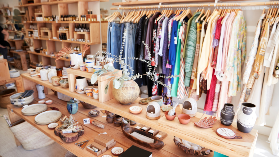 A gift shop with a variety of pottery and other small items on a shelf in the foreground and a clothing rack in the background.