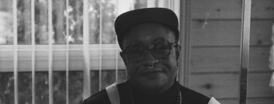 A black and white photograph of mel, a Black, agender, chubby person with medium brown skin. mel looks at the camera with a small smile.