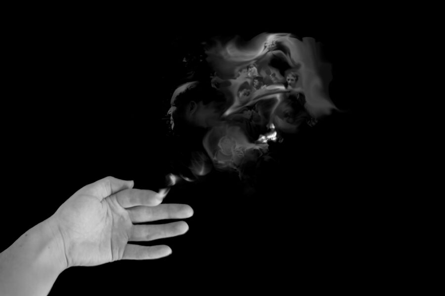 Black and white photo of a hand with a burning fingertip. In the smoke from the fingertip are many ghostly faces.