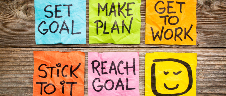 Six different coloured sticky notes on a wooden background with black block letters that say, Set goal, Make pan, Get to work, Stick to it, Reach goal and a smiley face.