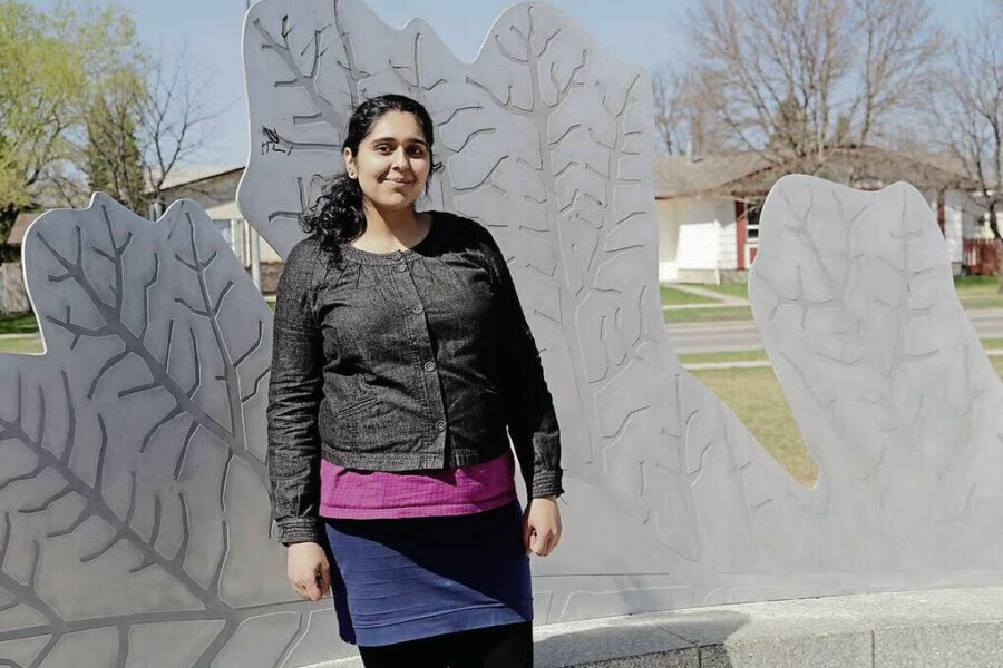 Photo of Gurpreet Sehra standing outdoors in front of a metal leaf sculpture. Sehra is a woman with light brown skin, long wavy black hair in a ponytail, wearing a black jacket, pink shirt and blue skirt.