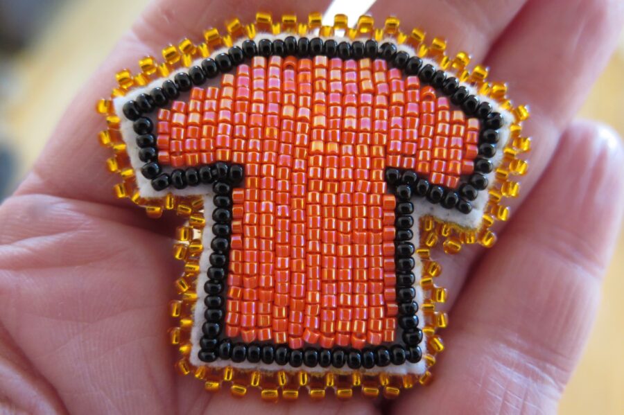 A beaded pin of an orange shirt outlined in black beads with a gold trim sits in the palm of a person's hand.