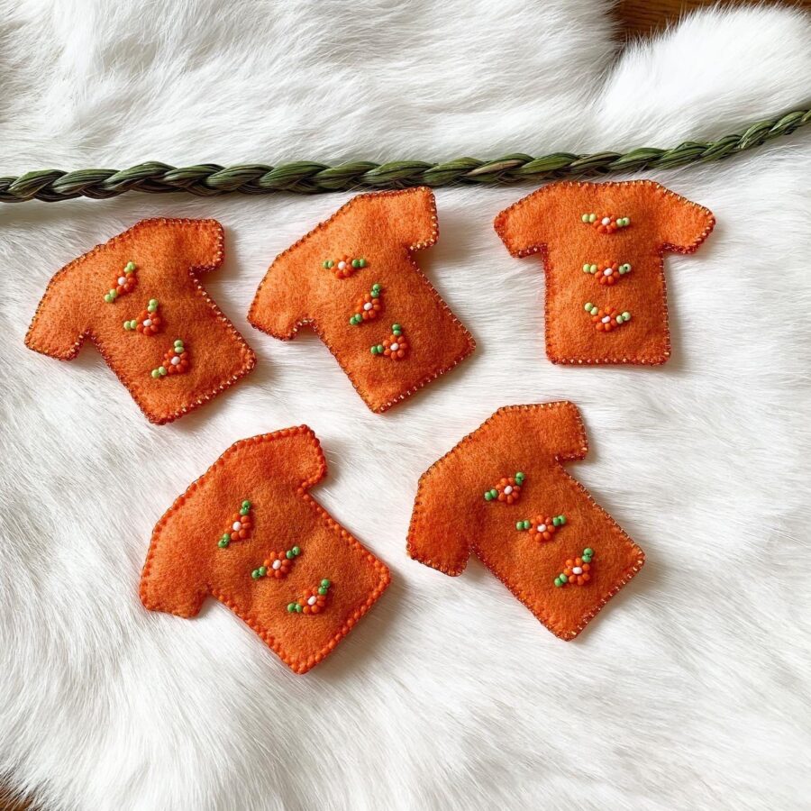 Five pins shaped like small t-shirts made of orange felt and beads lay on white fur with a braid of sweetgrass above.