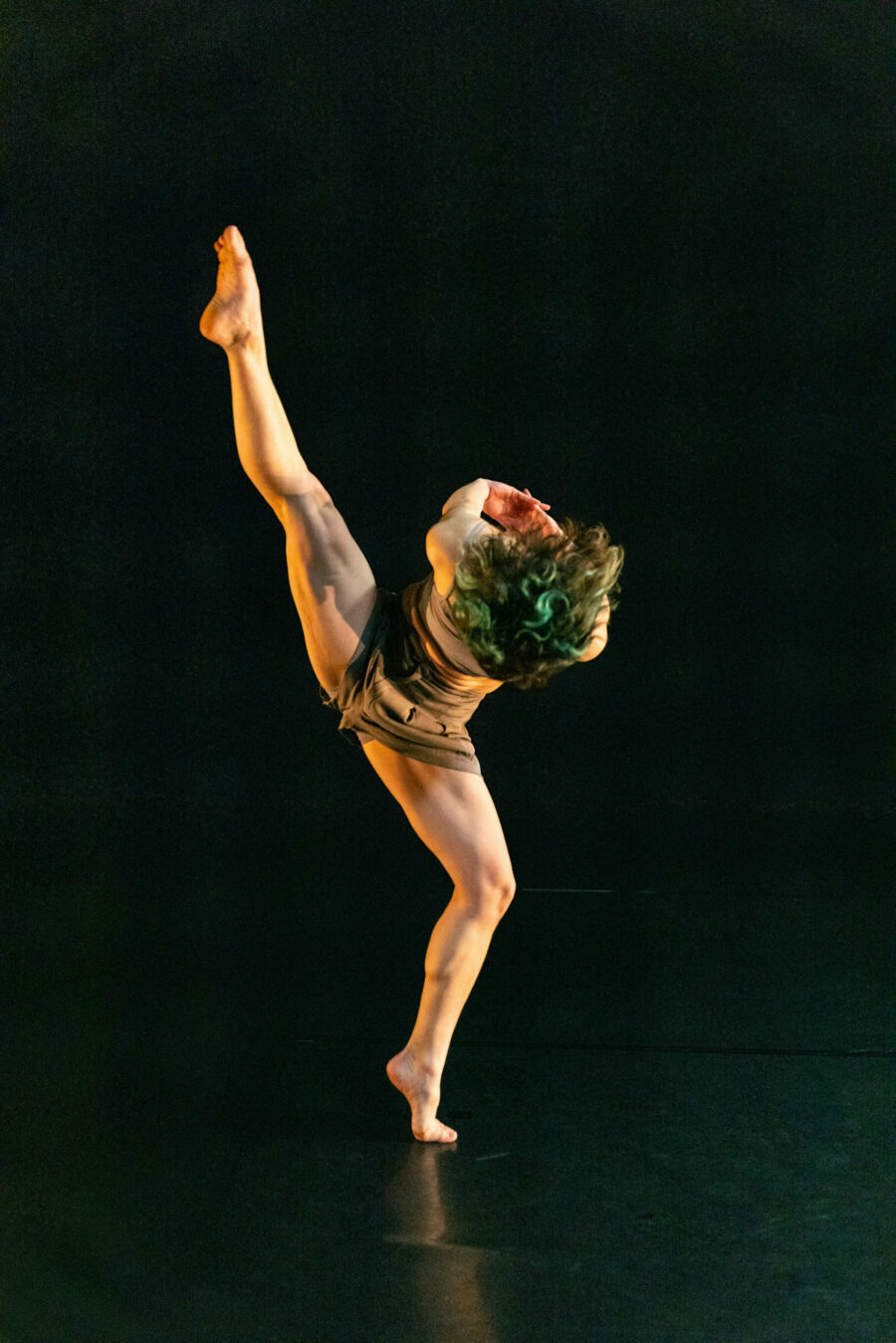 Carol-Ann Bohrn dancing in a darkened space, one leg raised in the air, leaning forward with interlaced fingers behind back.