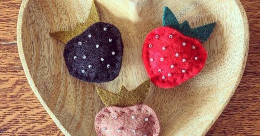 Three felt strawberries in dark purple, red and light pink with green tops and beads as seeds sit in a wooden heart shaped bowl.