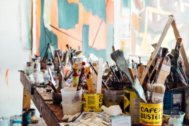 A messy wooden table in an artist's studio covered in paintbrushes and other painting supplies.