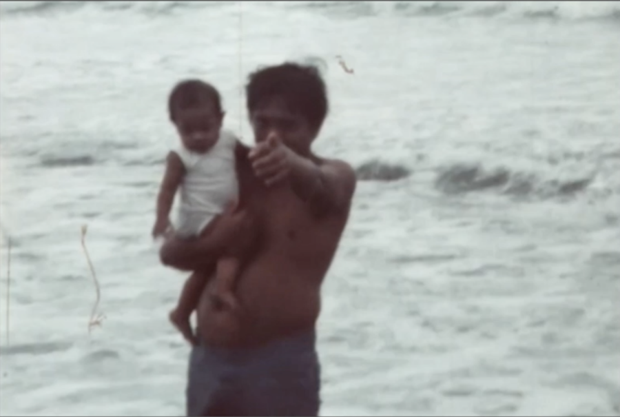 A still of a home film of a man at the beach holding a young child in one arm and pointing at the camera with the other.