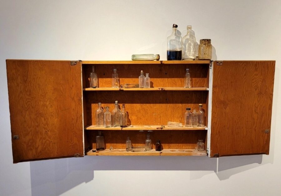 An open wooden medicine cabinet on a white wall with small glass medicine or liquor bottles inside.