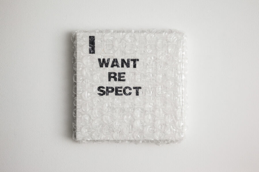 A square white canvas wrapped in bubble wrap hangs on a white wall. Black block letters say I WANT RE SPECT other letter outlined in light pencil are present but not legible.