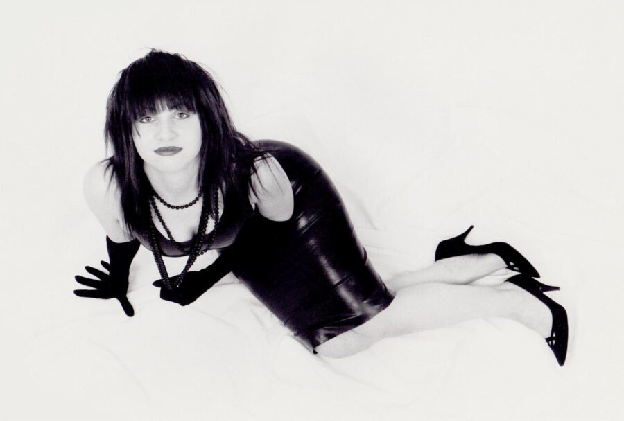 Black and white photo of Lydia Lunch wearing a black dress, high heels and gloves.  She is lying on a white background looking up at the camera.