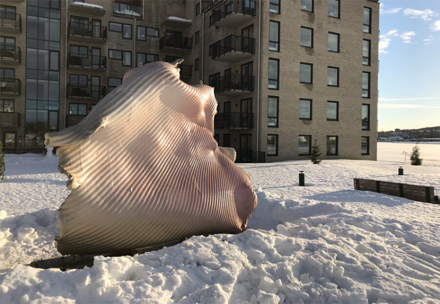 A large sculpture made of a sheet of metal formed into a wavy somewhat organic shape sits outside in the snow.