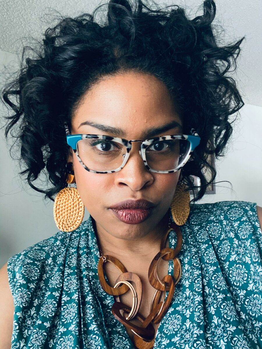 Headshot of Allison Yearwood. She is a black woman with curly black hair wearing a teal patterned top, leopard print glasses and large disc shaped earrings.