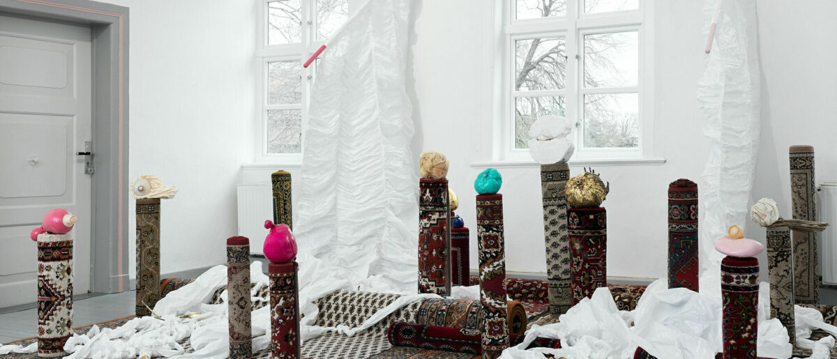 Art installation by Olga Ulmann, featuring rolled up area rugs set up like pillars with figurines perching on top of them and white material interspersed between the pillars.