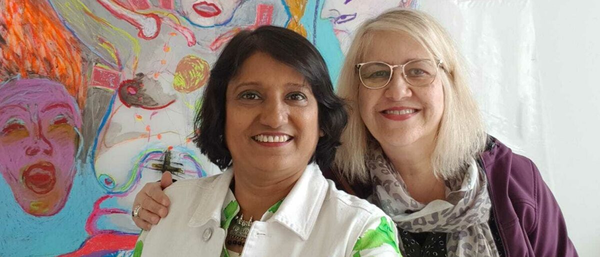 Artists Mahfuza Begum and Leesa Streifler pose smiling in front of a colourful painting.