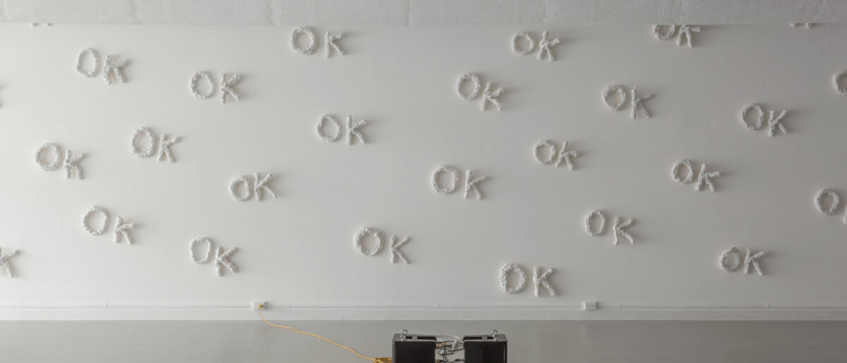 A photo in a gallery of an installation artwork by Tara Lynn MacDougall. The word OK in 3-D sculpted out of white material is repeated over and over on a white wall. On the floor are two speakers.
