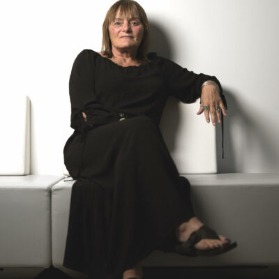 Photo of Liz Barron dressed in black sitting on a white chair in front of a white wall.