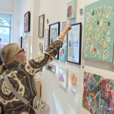 Two visitors to the 2023 MAWA members' show stand looking at the art. One points to an artwork on the wall.