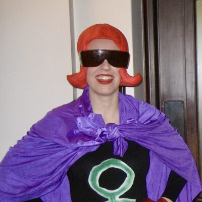 A woman in a red wig and sunglasses with her hands on her hips wearing tights a black body suit with a purple cape.