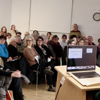 Patricia Bovey stands at a lectern in front of a crowd of 60 people seated in MAWA.