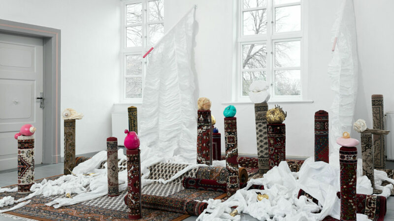 Art installation by Olga Ulmann, featuring rolled up area rugs set up like pillars with figurines perching on top of them and white material interspersed between the pillars.