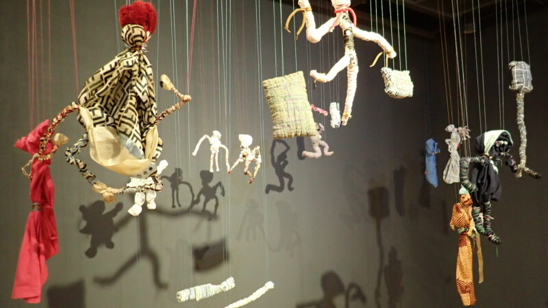 A variety of cloth dolls made out of old clothes hang from lines from the ceiling in a gallery.