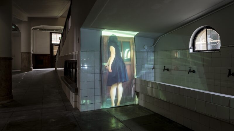 Photo of a video installation by Tomoko Inagaki. A video of a woman reaching to open a door is projected on a closed door. The closed door is underneath a staircase in a building with white tiled floors and walls.