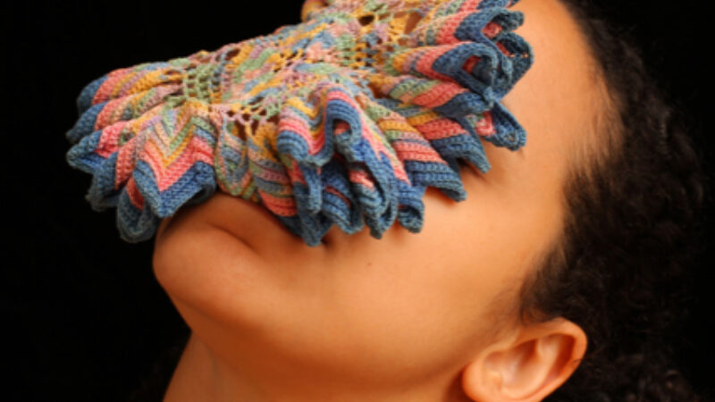 A circular, multicoloured crocheted piece sits on a person's face while they tip back their head.