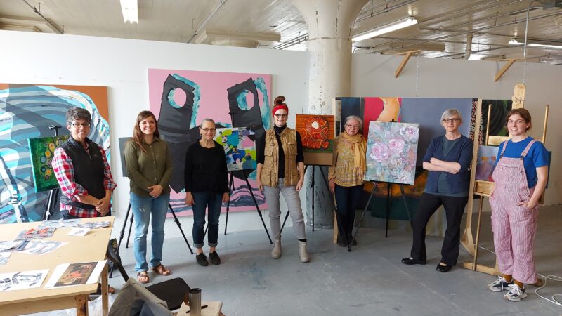 Seven people stand in a painter's studio.