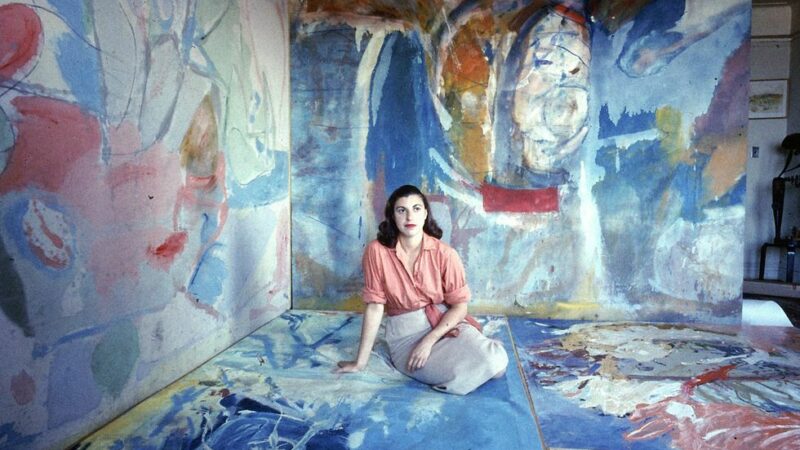 Artist, Helen Frankenthaler sits on the floor of her studio that is covered in paintings on the walls and the floor.