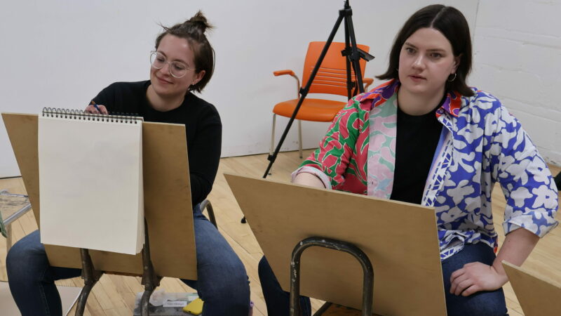 Two people sit on drawing horses in front of drawing boards and sketch.