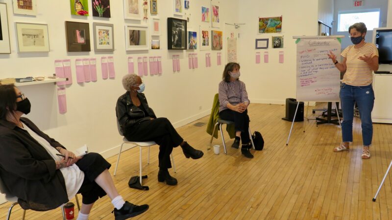 Three people sit in chairs in the MAWA space looking at Shawna Dempsey who is standing and talking next to a flip chart.