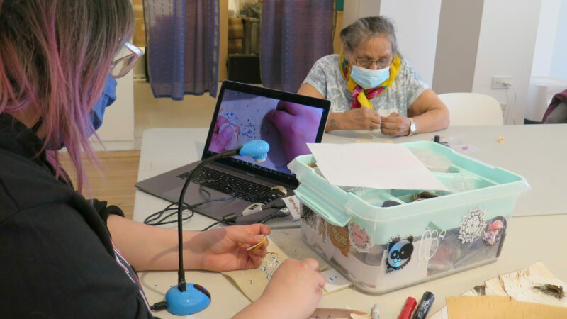 A photo of two people working on beading projects at a table. One person is using a laptop and webcam to share their work over Zoom.