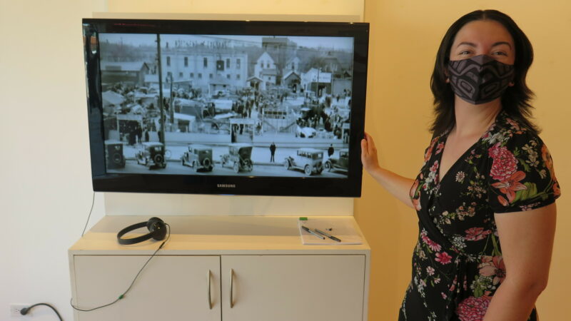 Sarah Paradis in a floral print dress and wearing a black mask, stands next to a television showing a black and white archival image of Winnipeg.