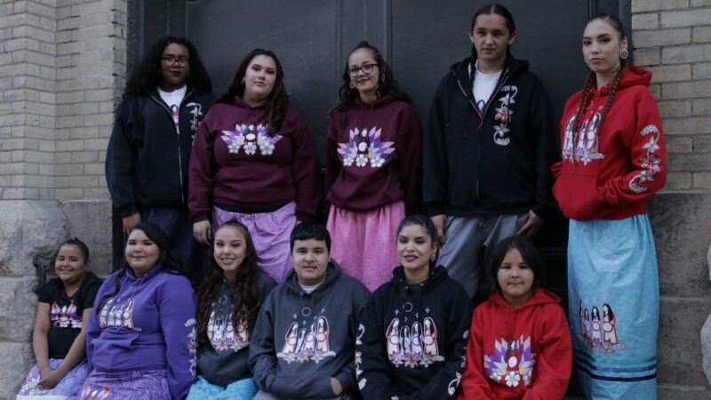 11 young people wearing hoodies and ribbon skirts or pants with designs by artist Christine Slater.