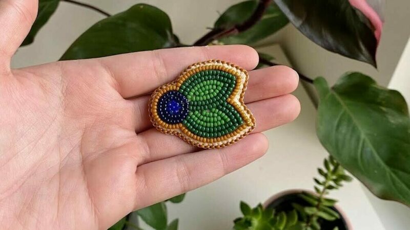 A beaded pin of a blueberry and two leaves lays in a person's hand.